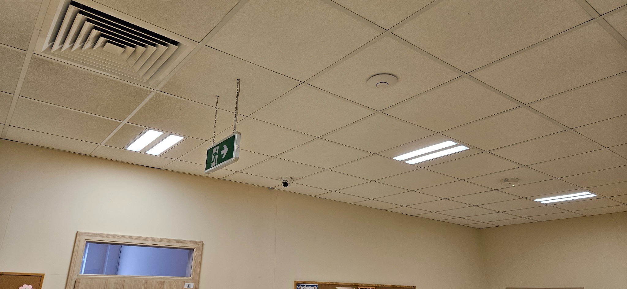 Kamera monitoringu na suficie podwieszanym z kratownicą, obok znak ewakuacyjny na łańcuszku, oświetlenie rastrowe i kratka wentylacyjna. Jasne ściany.