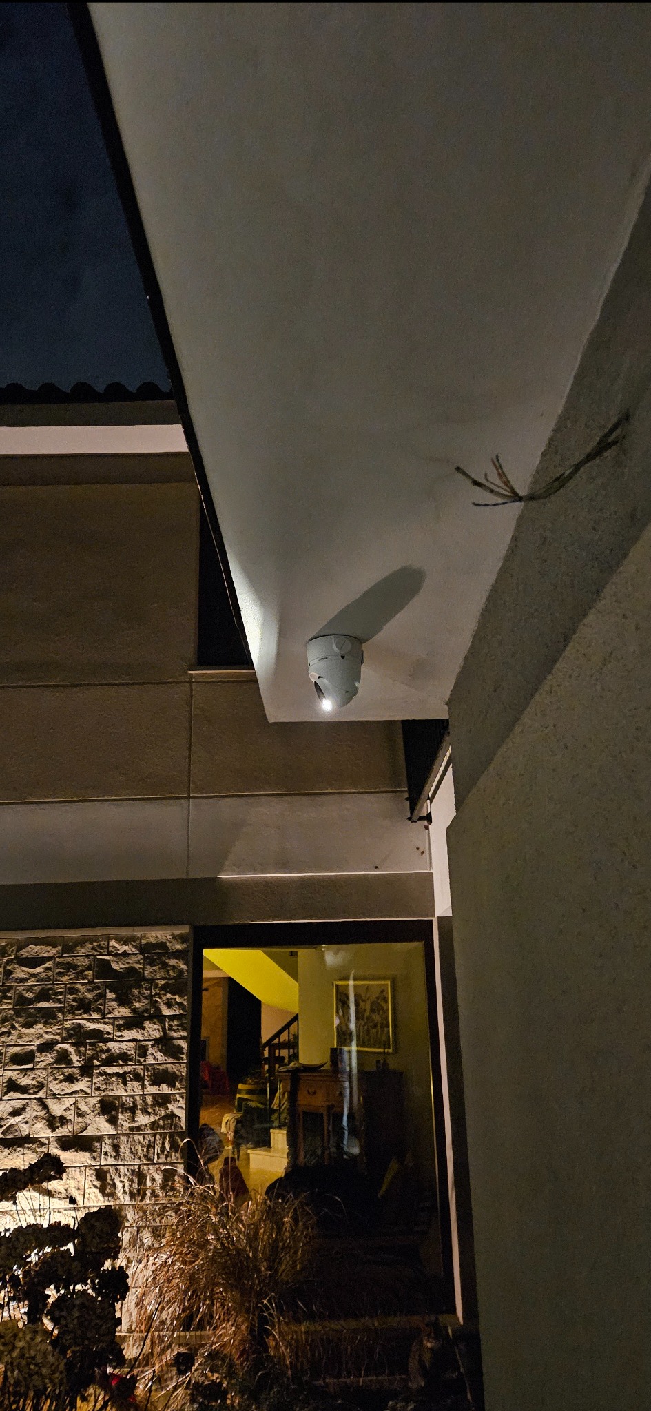 Kamera monitoringu zamontowana pod okapem budynku, widoczne przewody. Nocne oświetlenie, elewacja z kamienia. Widok na wnętrze domu przez okno.
