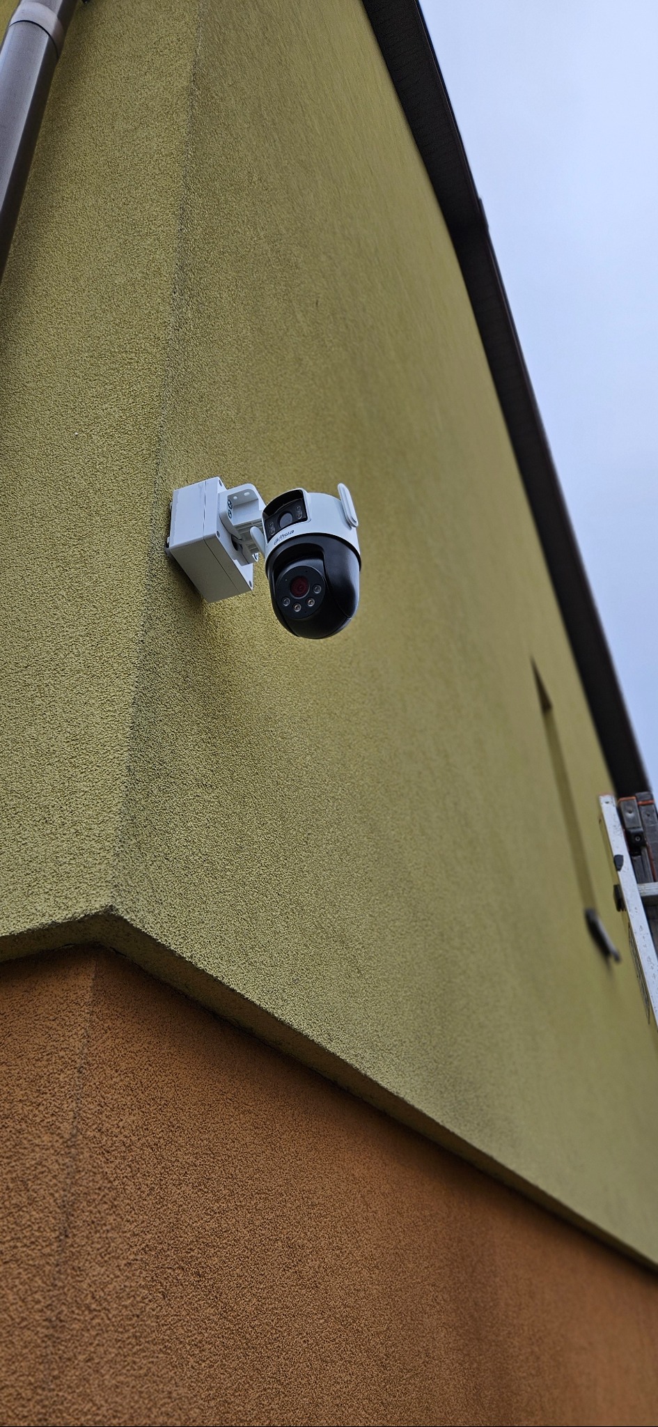 Kamera PTZ Dahua zamontowana na elewacji budynku o dwukolorowej strukturze, widoczna drabina oparta o ścianę. Ujęcie z dołu, pochmurne niebo w tle.