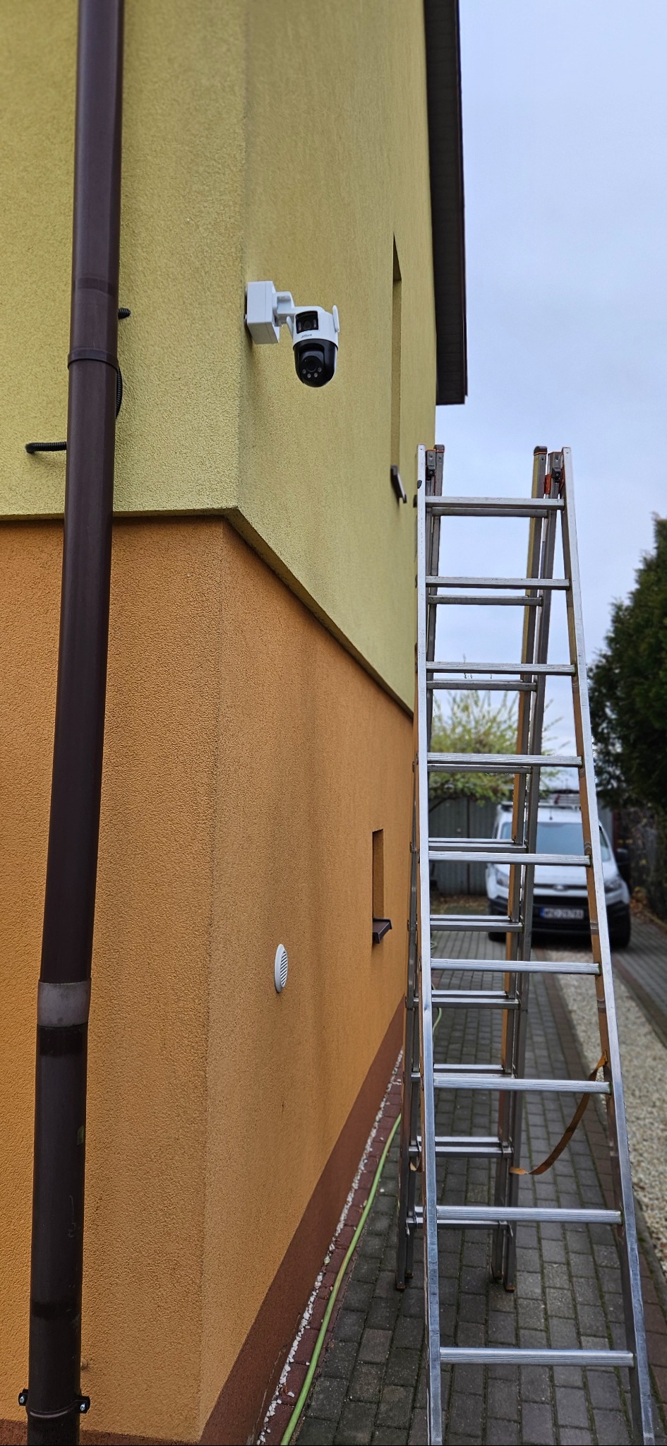 Kamera IP zamontowana na elewacji dwukondygnacyjnego budynku z drabiny. Widoczny fragment rynny spustowej i okna. W tle zaparkowany biały samochód dostawczy.