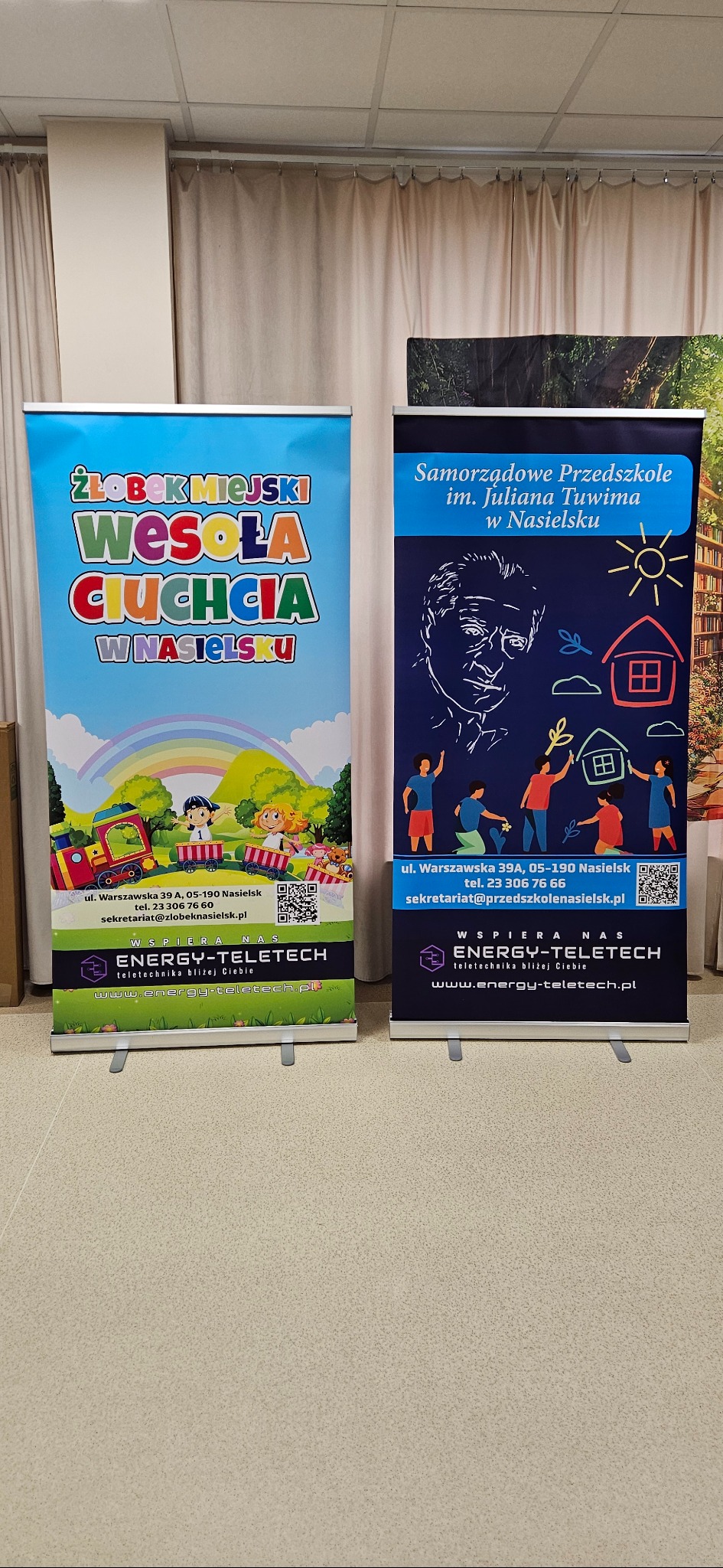 Dwa roll-upy reklamowe: kolorowy dla żłobka i granatowy dla przedszkola, oba z logo Energy-Teletech, stoją obok siebie na jasnym tle. Widoczne adresy i QR kody.