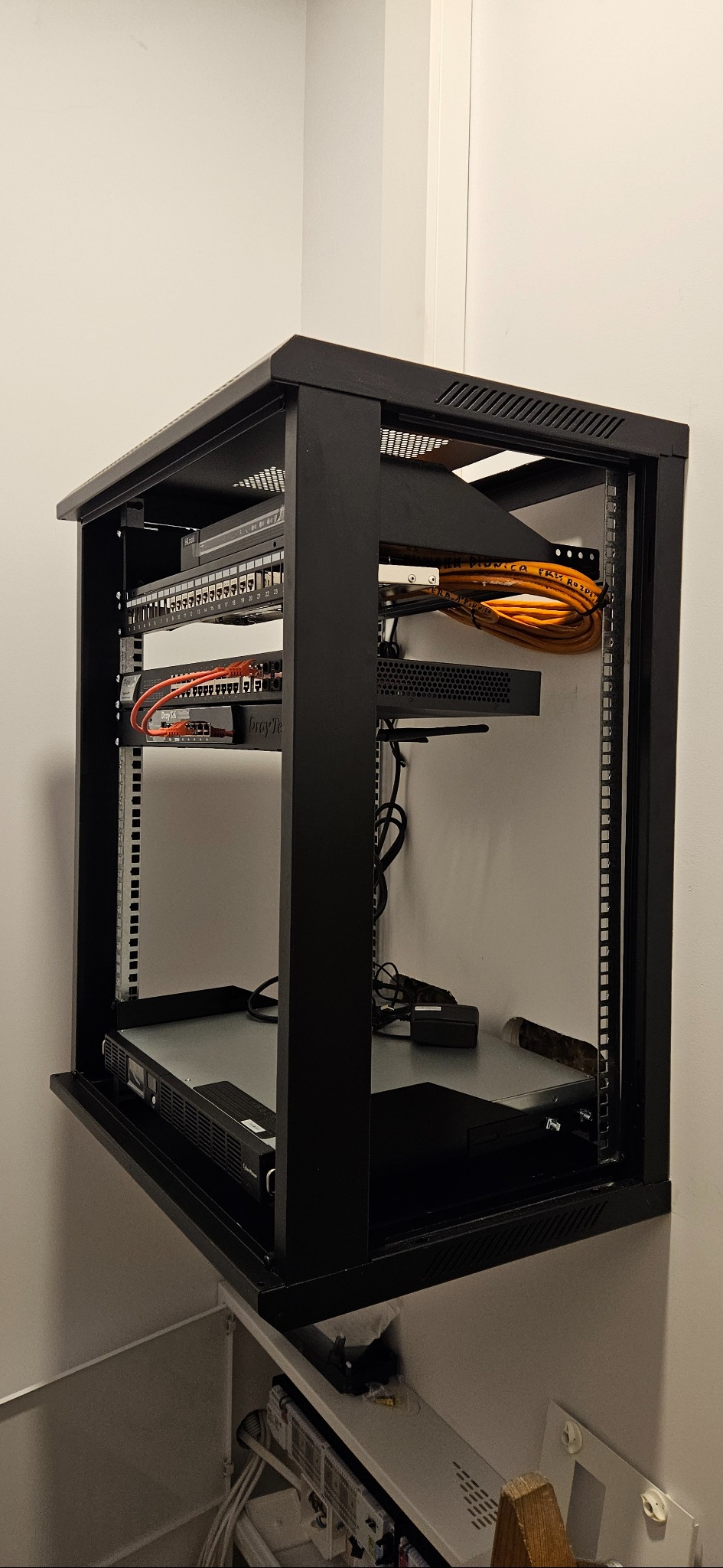 Ścienny, czarny rack teleinformatyczny z zamontowanymi urządzeniami sieciowymi, w tym switchem i routerem. Widoczne okablowanie i listwa zasilająca. Całość zamontowana w pomieszczeniu.