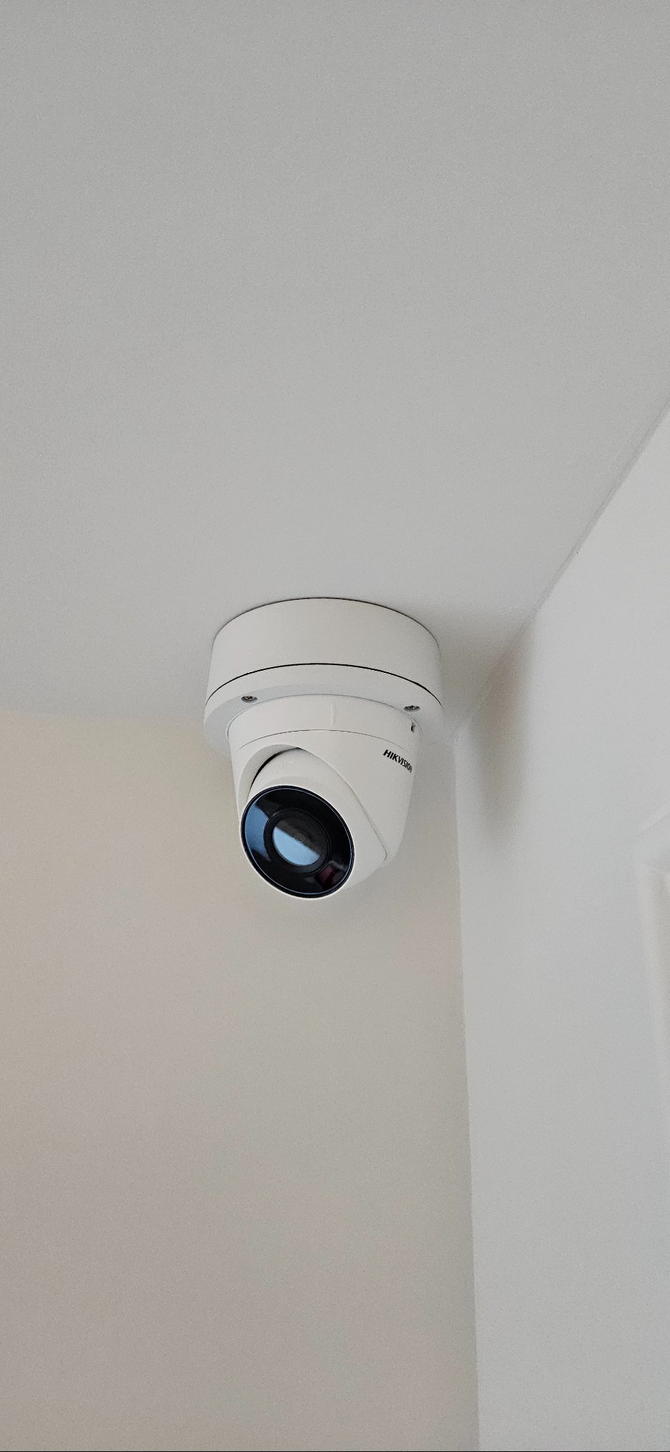 Kopułkowa kamera Hikvision zamontowana w rogu pomieszczenia z białymi ścianami i sufitem. Widoczny obiektyw i logo producenta. Ujęcie z dołu.