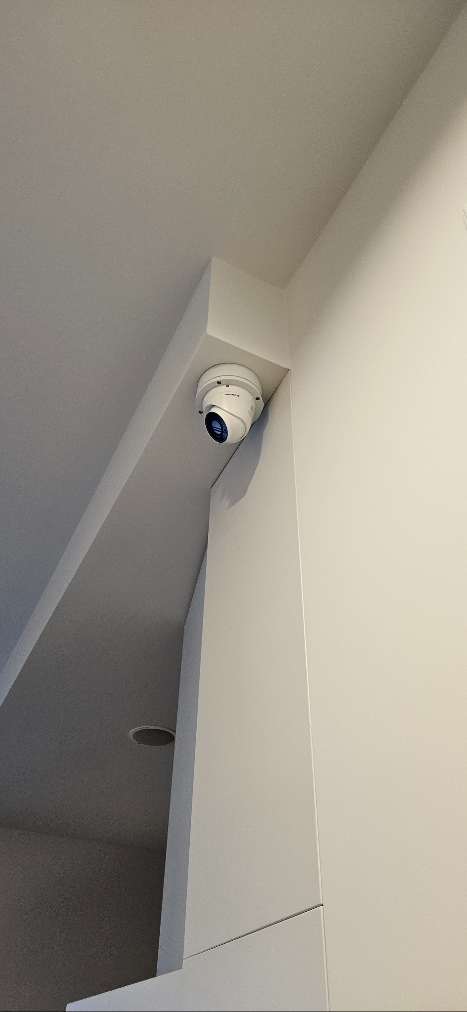 Kamera kopułkowa Hikvision zamontowana w rogu pomieszczenia, widok z dołu. Biała obudowa kamery kontrastuje z jasnym tłem ściany i sufitu.