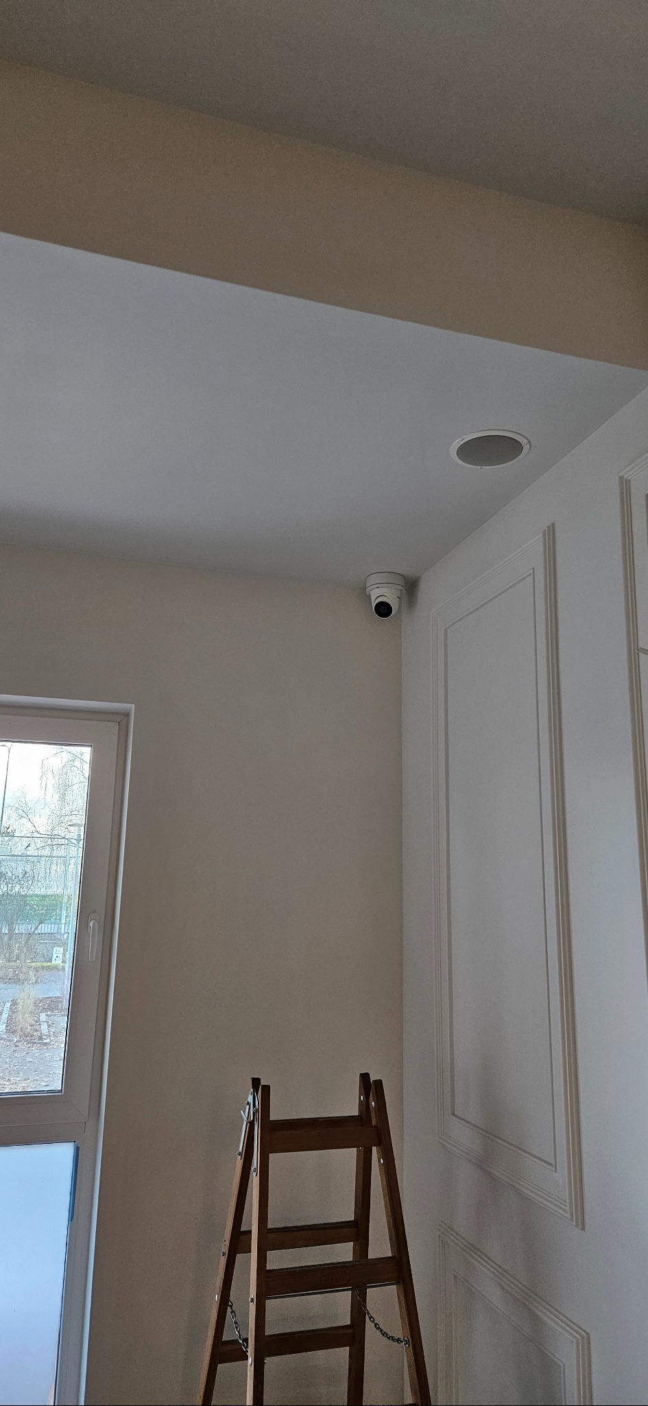 Kamera monitoringu zamontowana pod sufitem w pomieszczeniu z drabiną. Widoczny fragment okna i ozdobna ściana. Ujęcie z dołu.