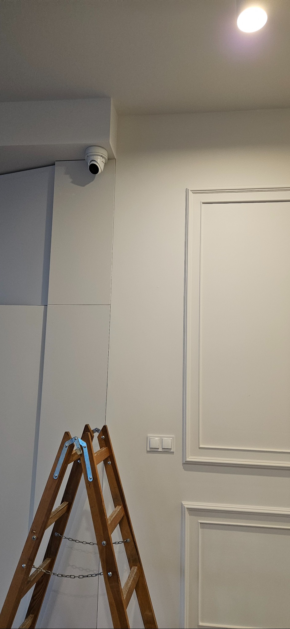Kamera monitoringu zamontowana pod sufitem w pomieszczeniu z dekoracyjną sztukaterią. Ustawiona drabina sugeruje trwający montaż lub serwis.