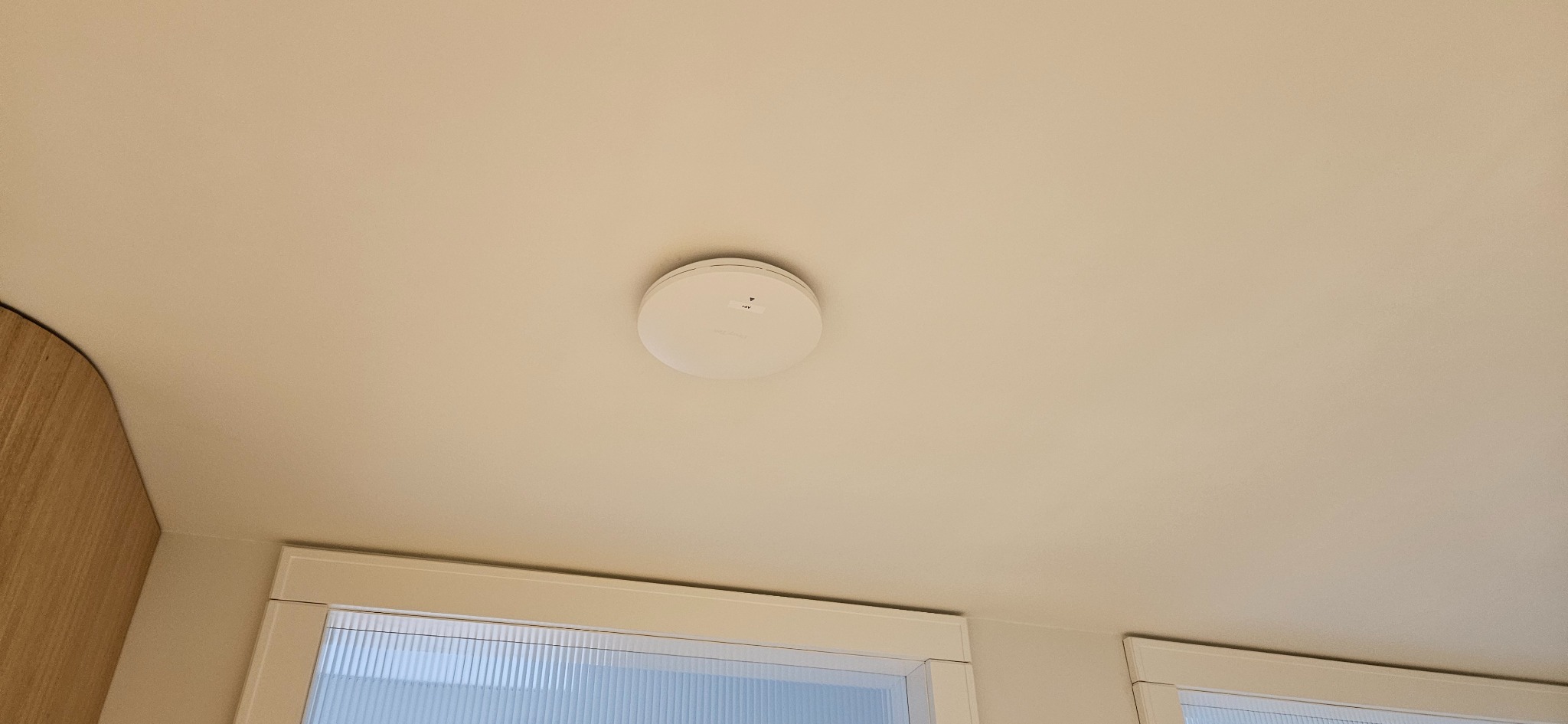Zamontowany, okrągły czujnik dymu na białym suficie w pomieszczeniu z oknem. Widoczny fragment drewnianej zabudowy. Ujęcie z dołu, centralnie.