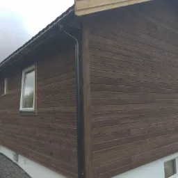 Elewacja domu z brązowymi deskami, biały cokół, czarna rynna, widoczne okna z białymi ramami i fragment drewnianego podestu z palet.