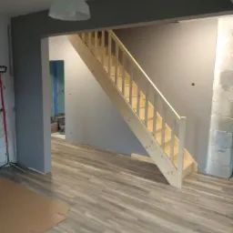 Surowe drewniane schody w nowo wykończonym wnętrzu z szarą ścianą i panelami podłogowymi, widoczne przygotowania do dalszych prac wykończeniowych.