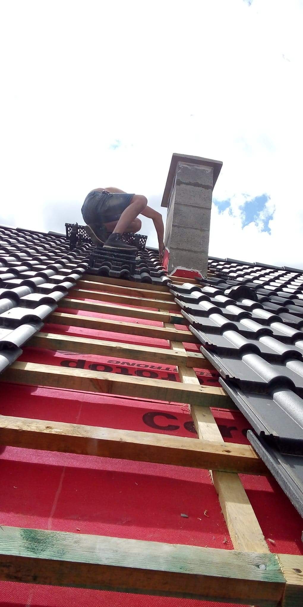 Montaż czarnej dachówki ceramicznej wokół komina na dachu w Pyrzycach, widoczne łaty i czerwona membrana dachowa. Prace dekarskie w toku.