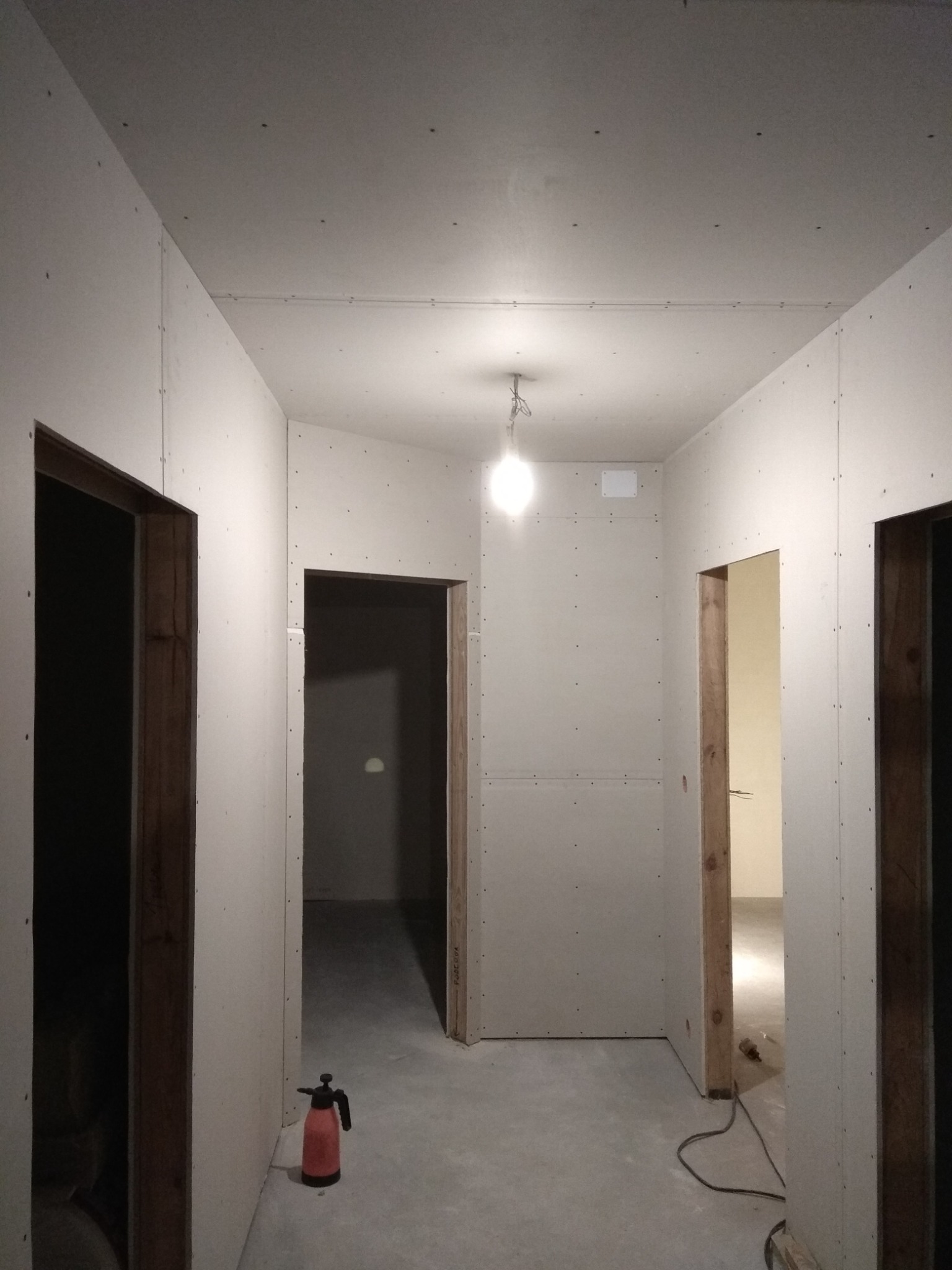 Korytarz w trakcie montażu płyt gipsowo-kartonowych na ścianach i suficie, widoczne profile, w tle żarówka na kablu i czerwony opryskiwacz ciśnieniowy.