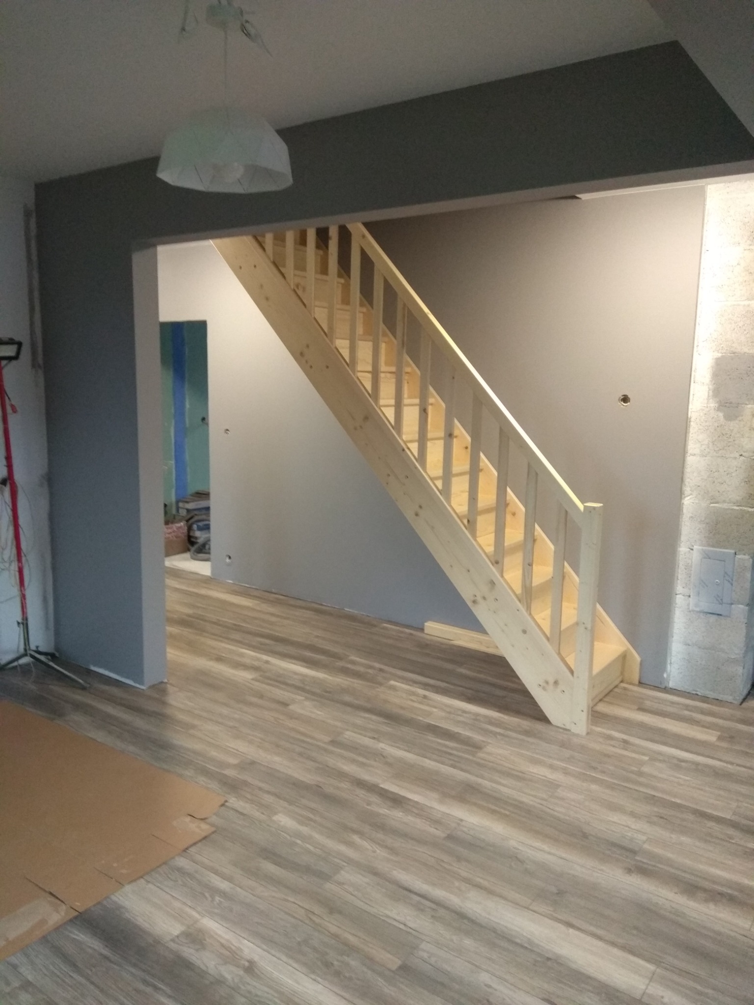 Surowe drewniane schody w nowo wykończonym wnętrzu z szarą ścianą i panelami podłogowymi, widoczne przygotowania do dalszych prac wykończeniowych.