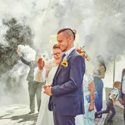 Uśmiechnięta para młoda w dymie z rac weselnych, panna młoda w okularach, pan młody w granatowym garniturze z przypiętym słonecznikiem, w tle goście weselni i dziecko biegnące po chodniku.