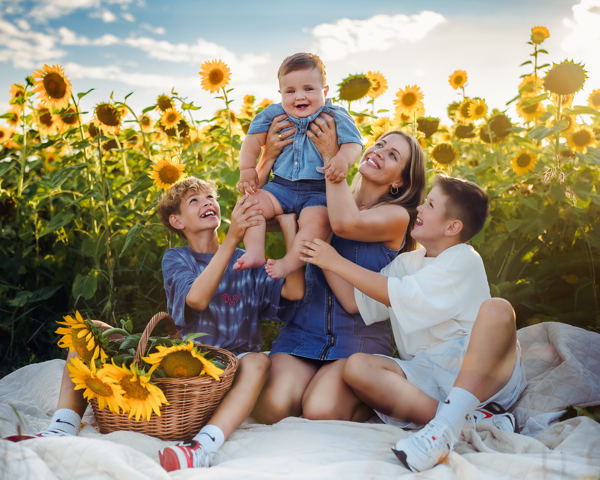 Uśmiechnięta mama trzymająca roześmianego niemowlaka nad głowami dwóch nastoletnich synów na tle pola słoneczników z koszem pełnym słoneczników, na białym kocu piknikowym.