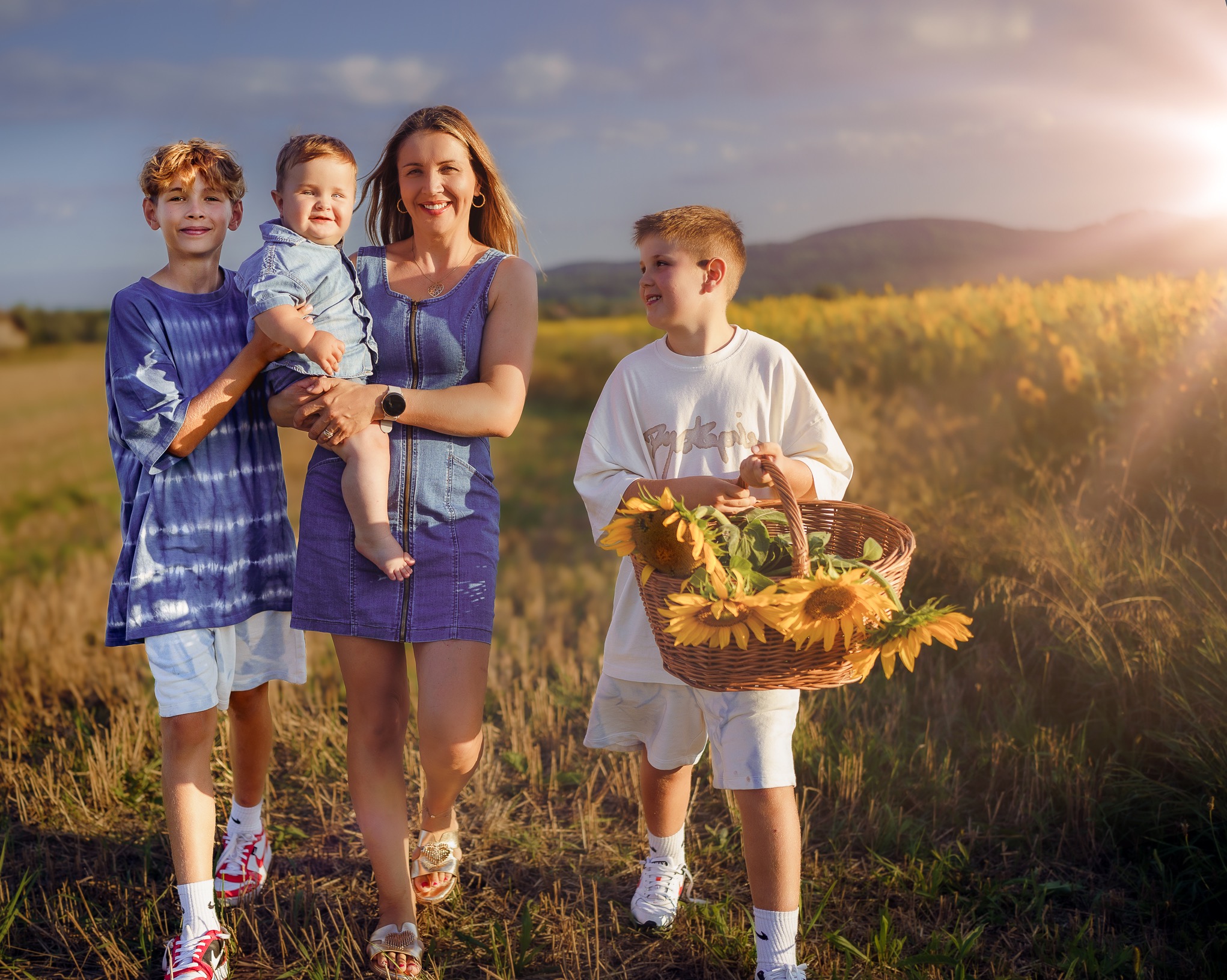 Uśmiechnięta matka z dwoma synami i małym dzieckiem na rękach, spacerująca po polu słoneczników w promieniach zachodzącego słońca. Jeden syn niesie koszyk pełen słoneczników.