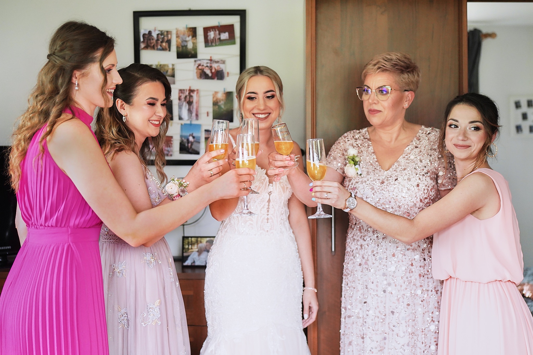 Panna młoda w koronkowej sukni i druhny w różowych sukienkach wznoszą toast szampanem, zdjęcia przypięte do tablicy w tle.