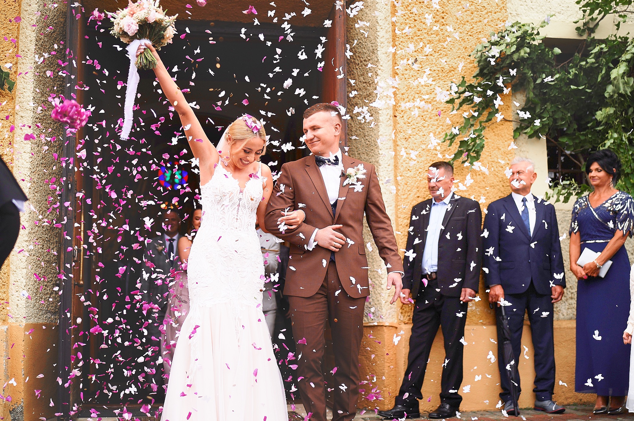 Uśmiechnięta para młoda wychodzi z kościoła, obsypywana konfetti w kształcie białych motyli i różowych płatków kwiatów, panna młoda z bukietem w uniesionej ręce, goście w tle.