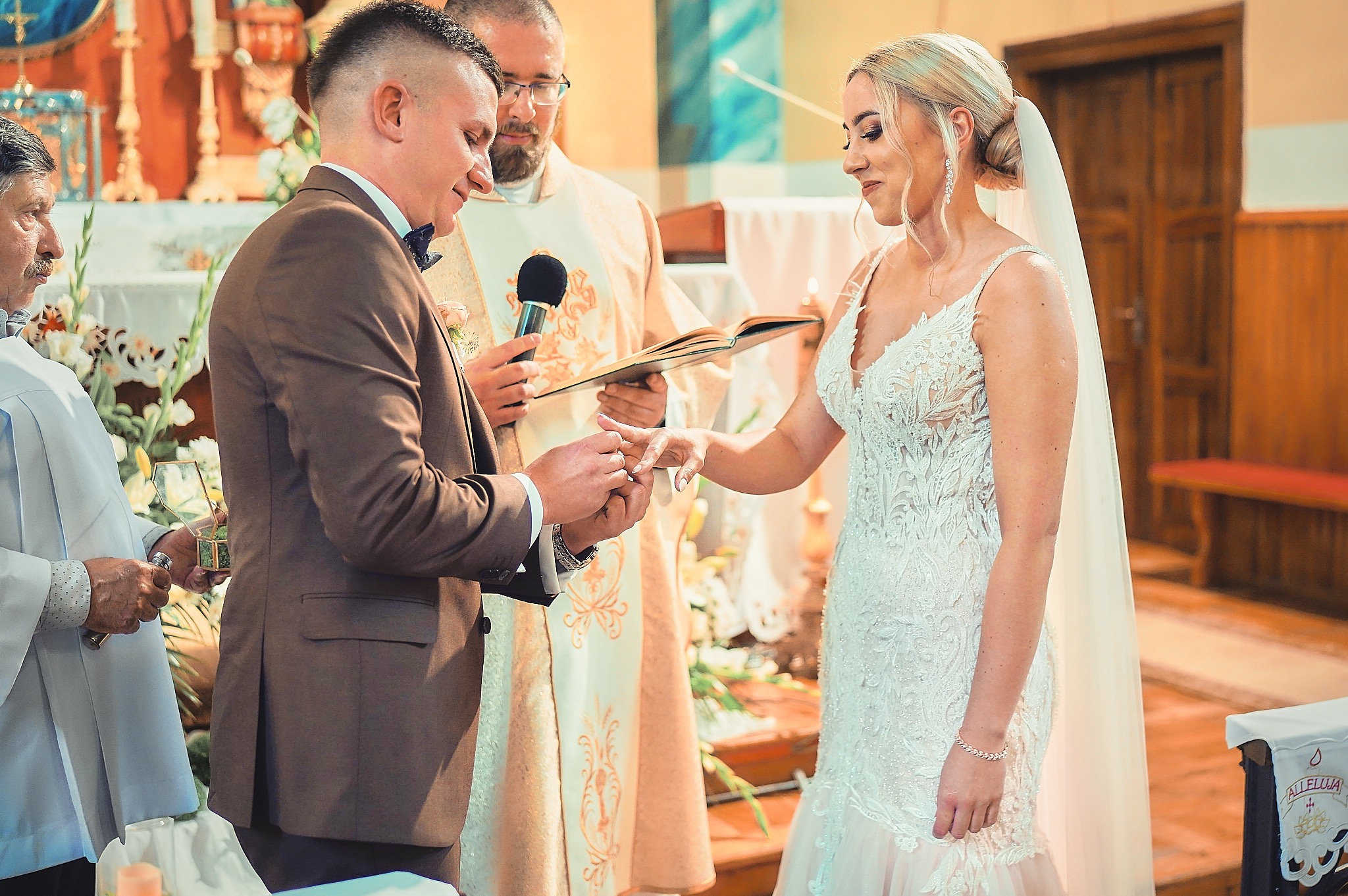 Uśmiechnięta panna młoda w koronkowej sukni ślubnej przyjmuje obrączkę od pana młodego w brązowym garniturze, w tle ksiądz i dekoracje kwiatowe w kościele.