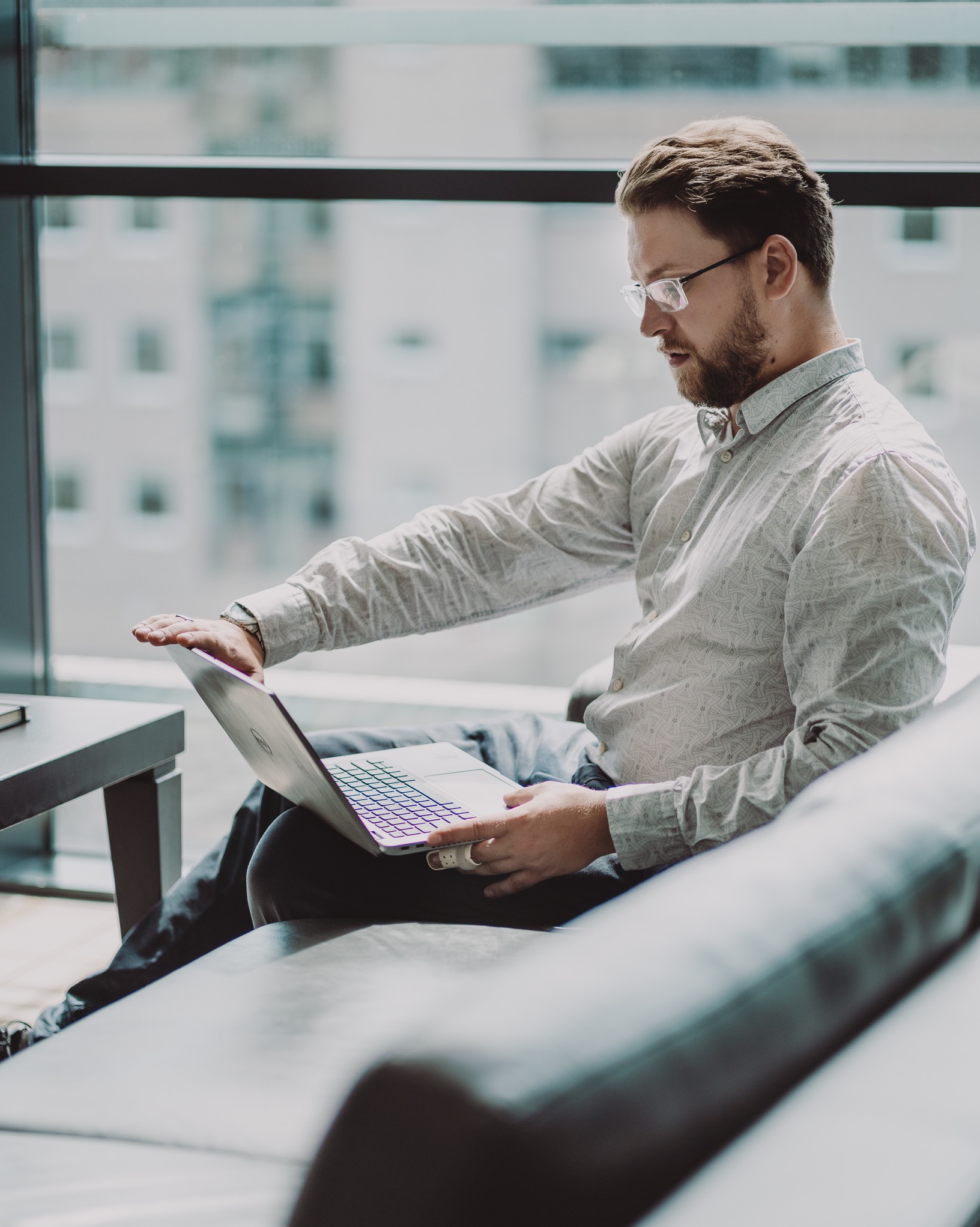 Mężczyzna w okularach siedzi na kanapie z laptopem w Wysokiej Strzyżowskiej, zamykając go jedną ręką. Widok przez okno na budynki.