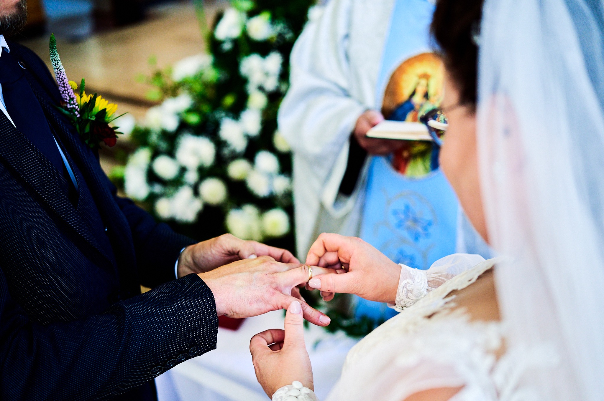 Zakładanie obrączki podczas ceremonii ślubnej, zbliżenie na dłonie pary młodej, w tle ksiądz z ikoną.