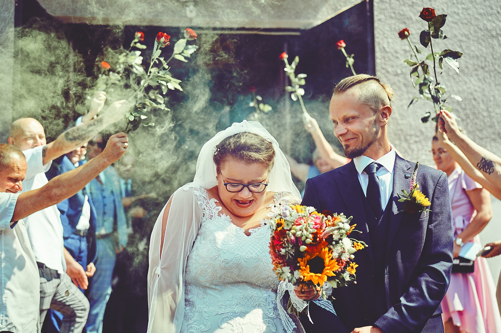 Uśmiechnięta panna młoda w koronkowej sukni i okularach, trzymająca bukiet słoneczników, obok pan młody w garniturze. W tle goście weselni rzucający płatki róż.