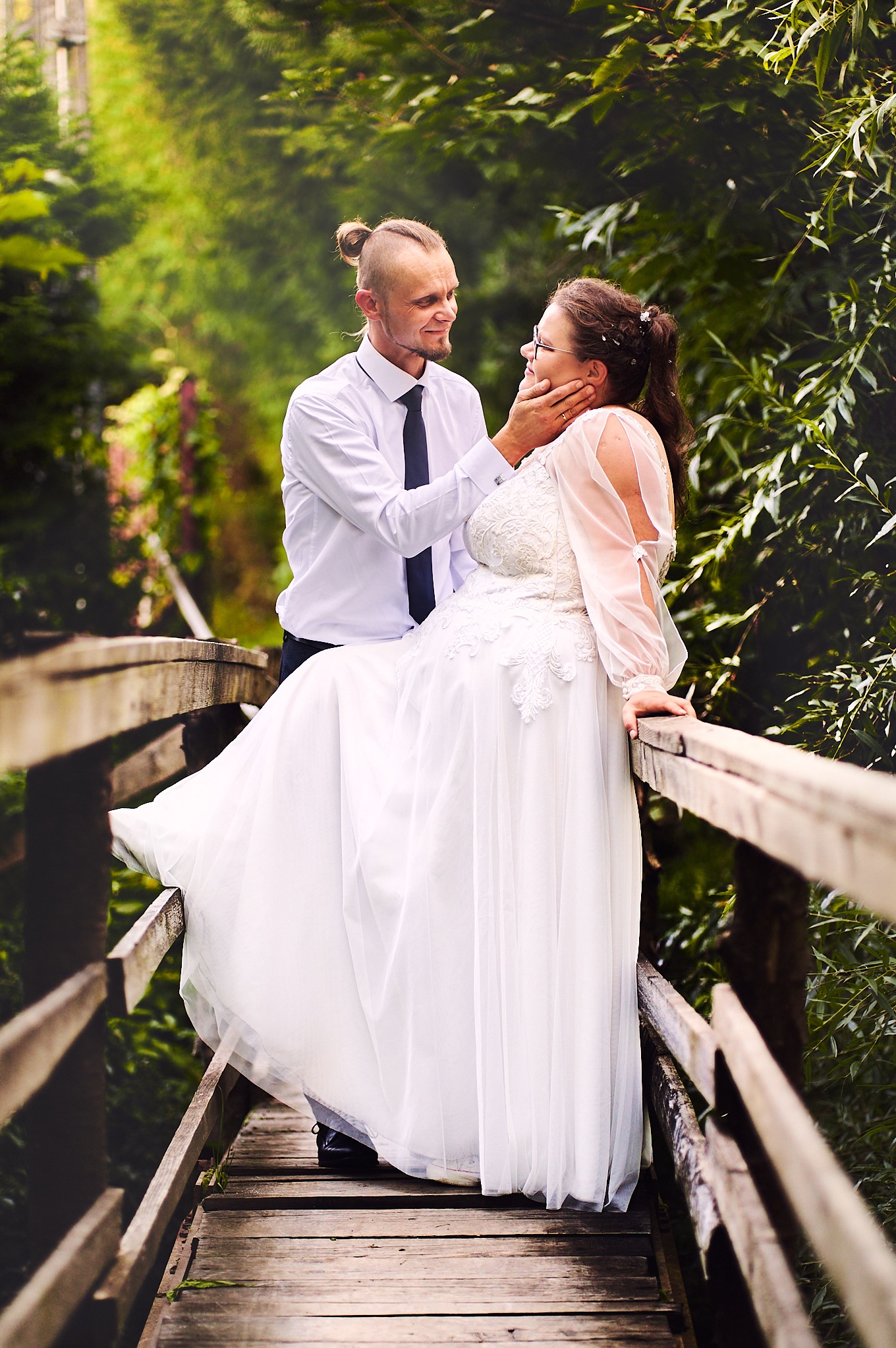 Uśmiechnięta para młoda na drewnianym moście wśród zieleni: on w białej koszuli i krawacie, ona w koronkowej sukni ślubnej z długimi, bufiastymi rękawami, delikatnie dotykana przez niego po twarzy.