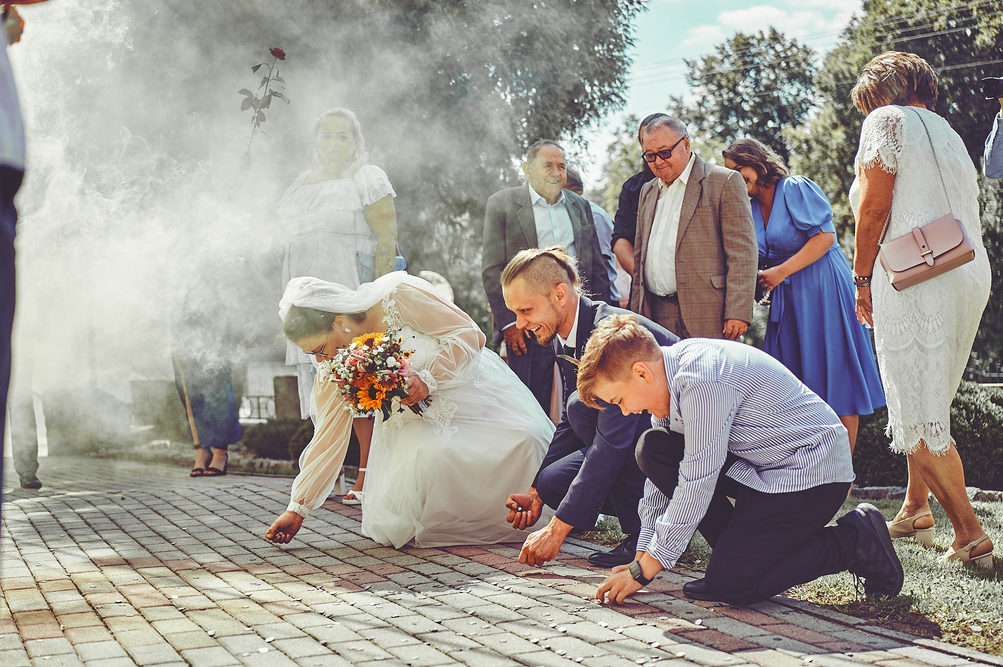 Para młoda i młody chłopak zbierają monety z brukowej ścieżki po ceremonii ślubnej, w tle goście weselni i dym.