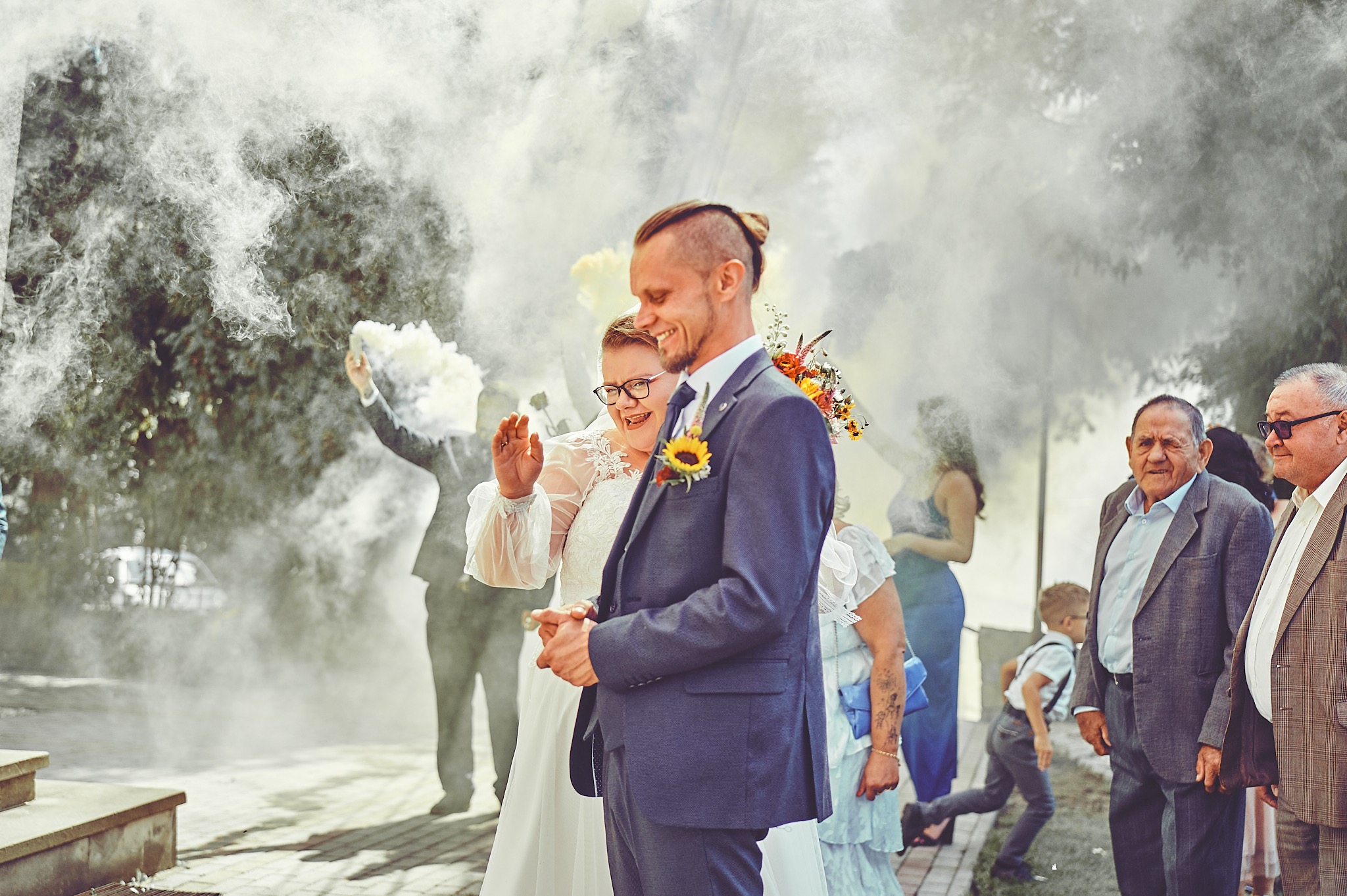 Uśmiechnięta para młoda w dymie z rac weselnych, panna młoda w okularach, pan młody w granatowym garniturze z przypiętym słonecznikiem, w tle goście weselni i dziecko biegnące po chodniku.
