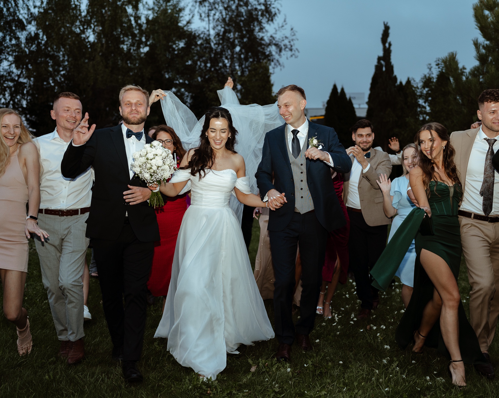Uśmiechnięta para młoda w otoczeniu gości weselnych, idąca po trawie. Panna młoda trzyma bukiet białych róż. W tle drzewa i wieczorne niebo.
