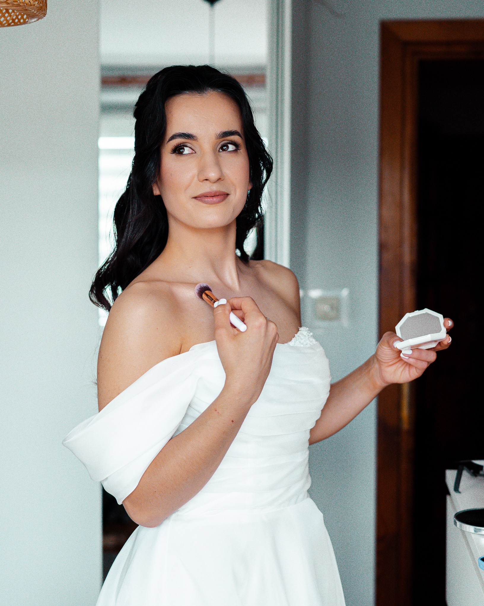 Panna młoda w białej sukni, z ciemnymi włosami, aplikuje makijaż pędzlem, spoglądając w bok. W tle widoczne drzwi i lustro.