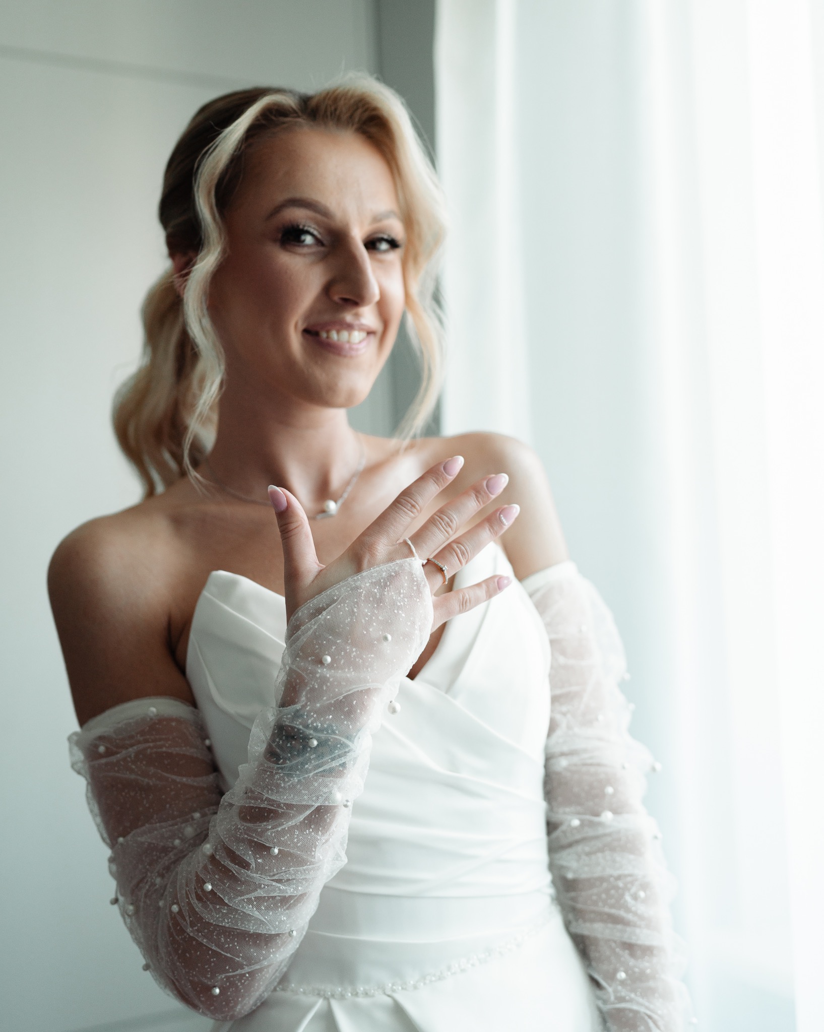 Uśmiechnięta panna młoda w białej sukni, z delikatnymi rękawiczkami z perełkami, prezentuje pierścionek zaręczynowy na tle jasnego okna.
