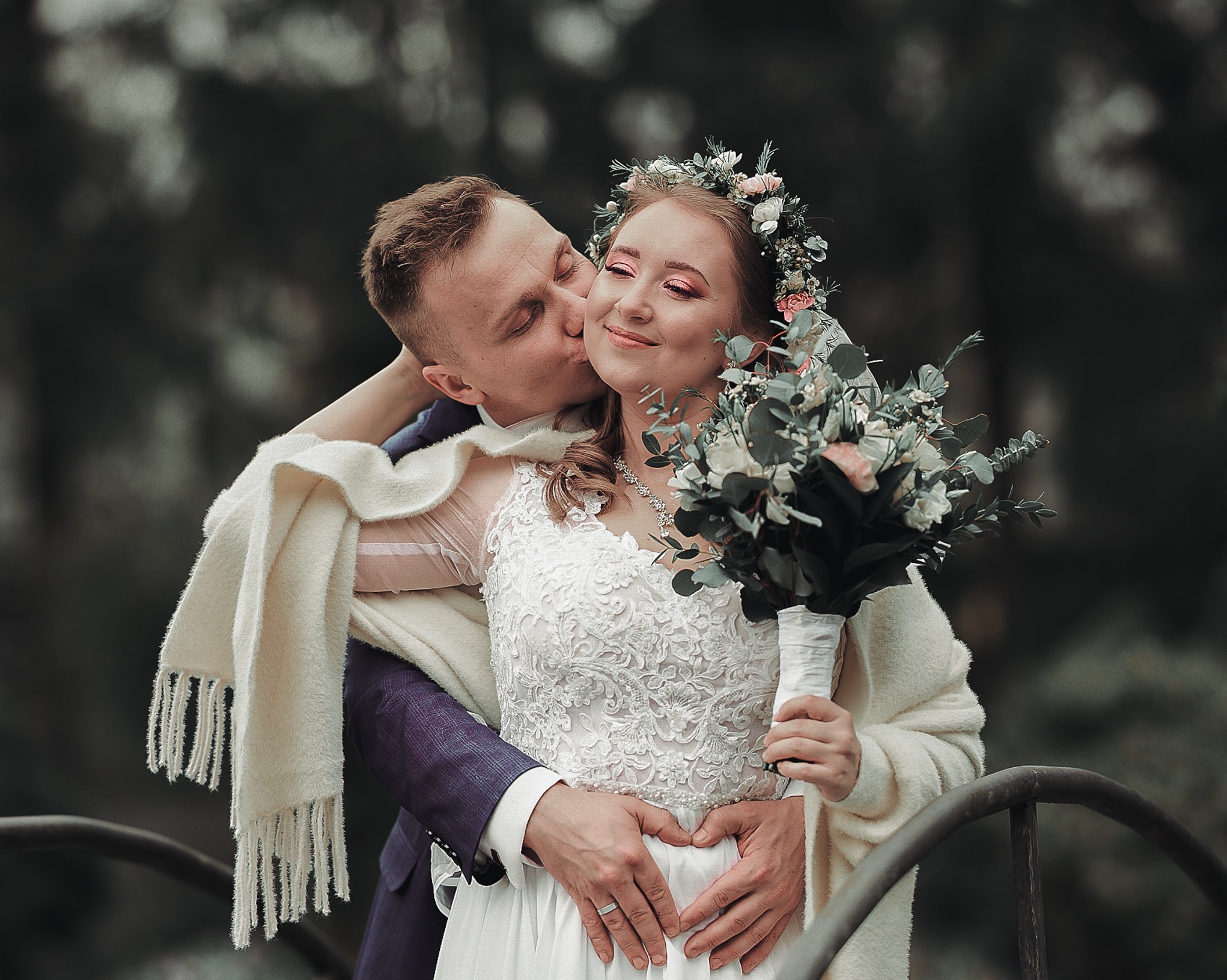Uśmiechnięta panna młoda w wianku i z bukietem, całowana w policzek przez pana młodego w fioletowym garniturze, otulona białym szalem na tle zieleni.