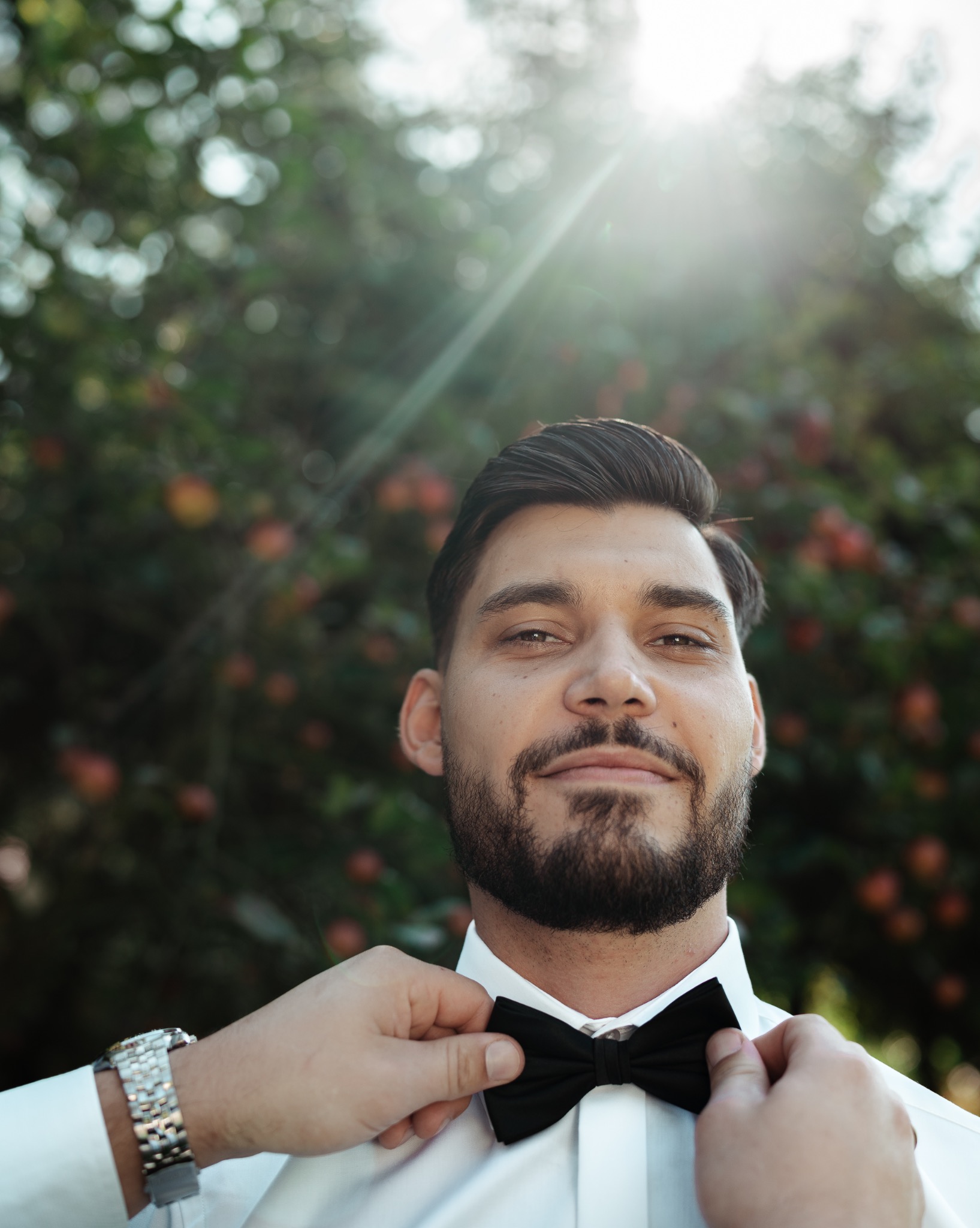 Uśmiechnięty mężczyzna z brodą poprawia czarną muchę na tle drzewa z owocami, w jasnym świetle słonecznym. Widoczne dłonie pomagające w poprawieniu muchy.