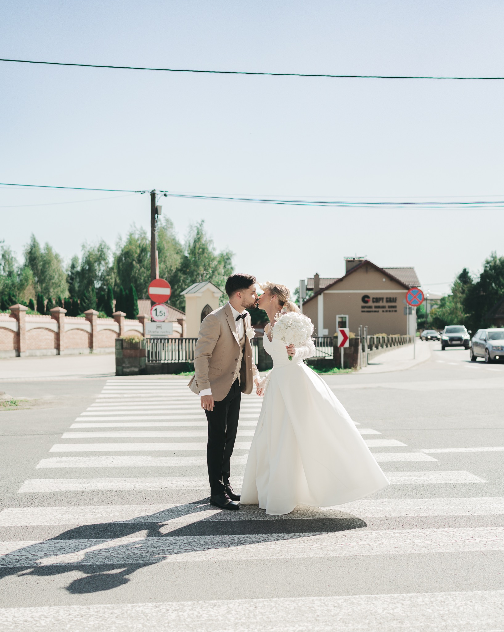 Para młoda w strojach ślubnych całuje się na przejściu dla pieszych. W tle budynki i zieleń. Jasne, naturalne światło.
