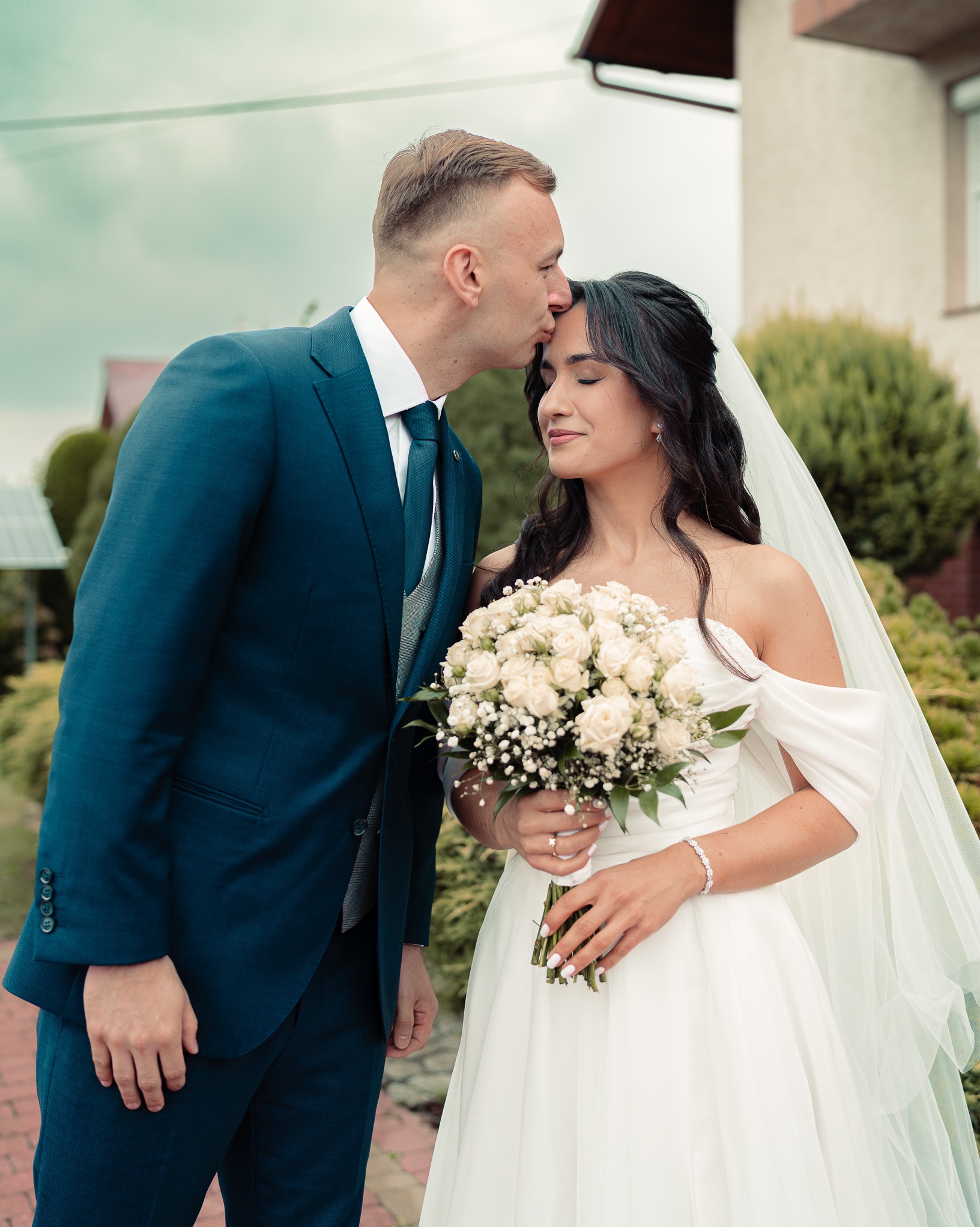 Pan młody w granatowym garniturze całuje pannę młodą w czoło. Panna młoda trzyma bukiet białych róż. W tle dom i zieleń. Romantyczny moment ślubny.