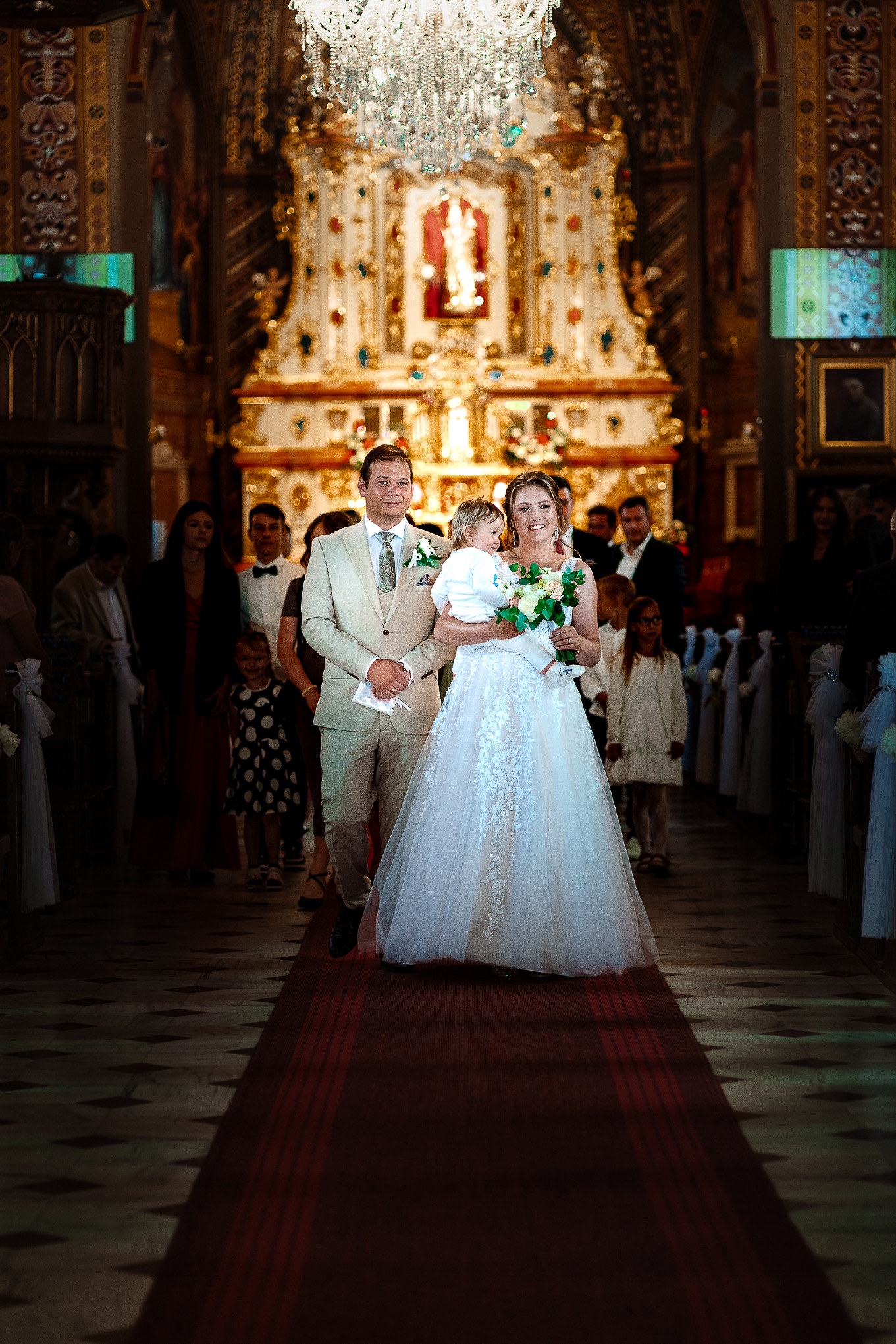Para młoda z dzieckiem idzie w kościele, suknia z koronką, bukiet kwiatów, elegancki garnitur, uroczysta atmosfera, bogato zdobione wnętrze, kryształowy żyrandol.