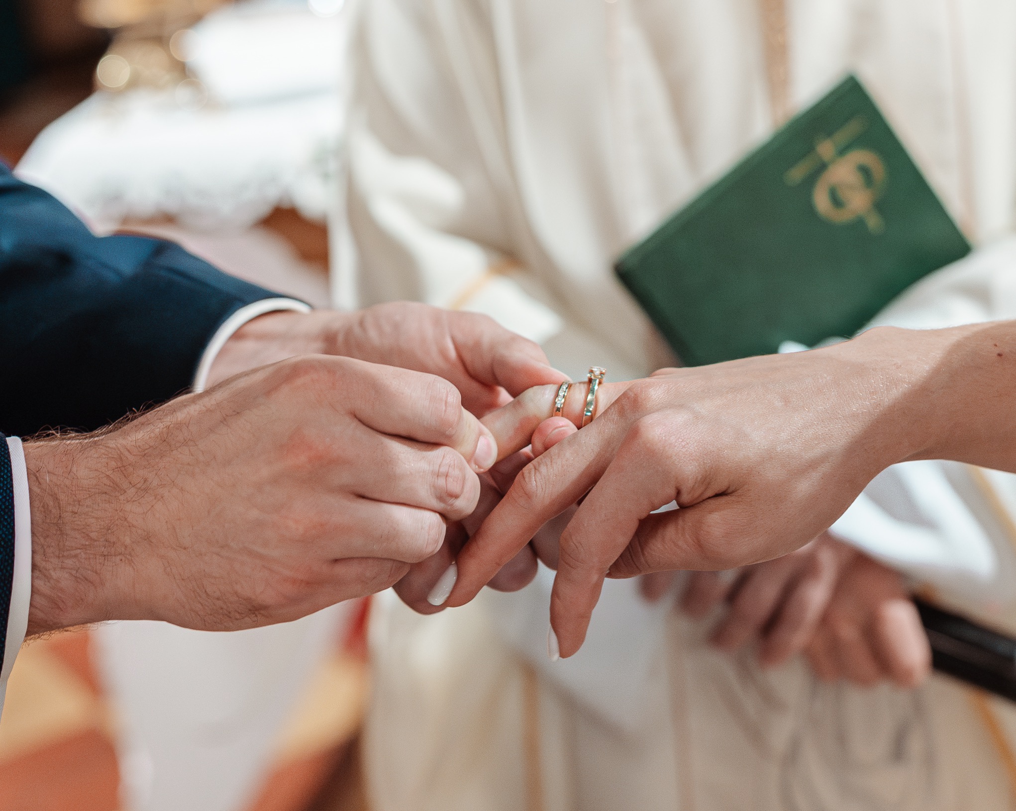 Zbliżenie dłoni nakładających złotą obrączkę na palec podczas ceremonii ślubnej, z zielonym modlitewnikiem w tle i delikatnym oświetleniem.