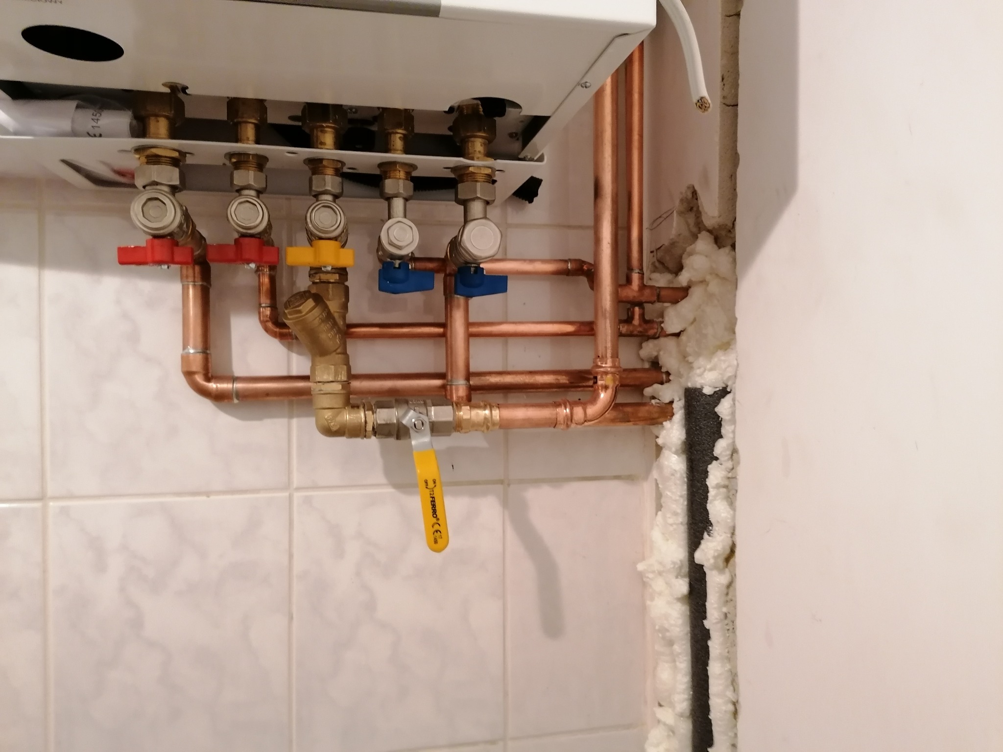 Nowa instalacja centralnego ogrzewania z miedzianymi rurami, zaworami z kolorowymi uchwytami (czerwony, żółty, niebieski) i kotłem, widoczne białe płytki na ścianie i pianka uszczelniająca wokół rur.