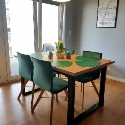 Stół z drewnianym blatem i czarnymi, metalowymi nogami w nowoczesnym wnętrzu, nakryty zielonymi podkładkami i ozdobiony kwiatami w doniczce oraz ceramicznymi figurkami.