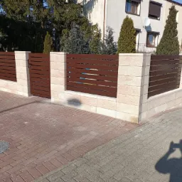 Nowoczesna, brązowa brama wjazdowa z poziomymi panelami, osadzona w jasnych, kamiennych słupkach i murku, na tle domu i zieleni.