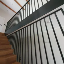 Nowoczesne, czarne poręcze schodowe z pionowymi szczeblami, zamontowane w domu w Śremie, widok z dołu na drewniane schody i białe ściany.