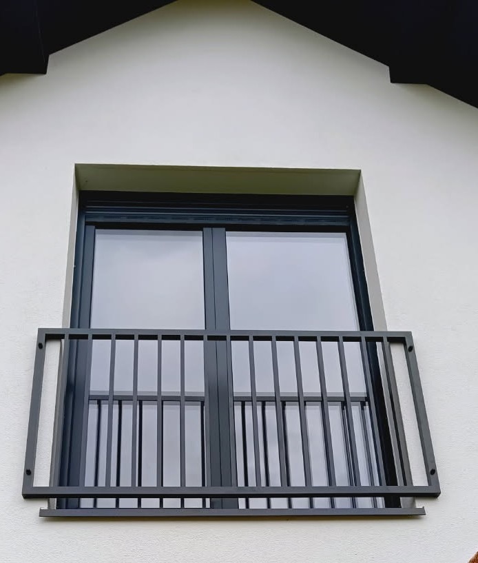 Zewnętrzna poręcz okienna z pionowymi szczeblami, zamontowana na białej elewacji budynku z czarną stolarką okienną.