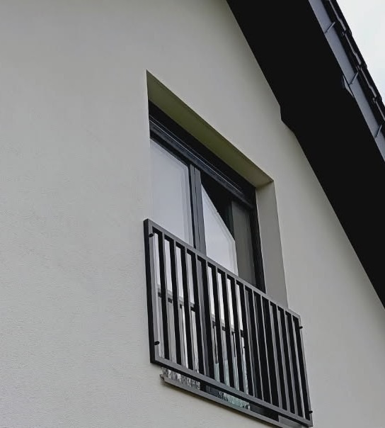 Balkon francuski z czarną, metalową balustradą, zamontowany na elewacji budynku z oknem balkonowym.