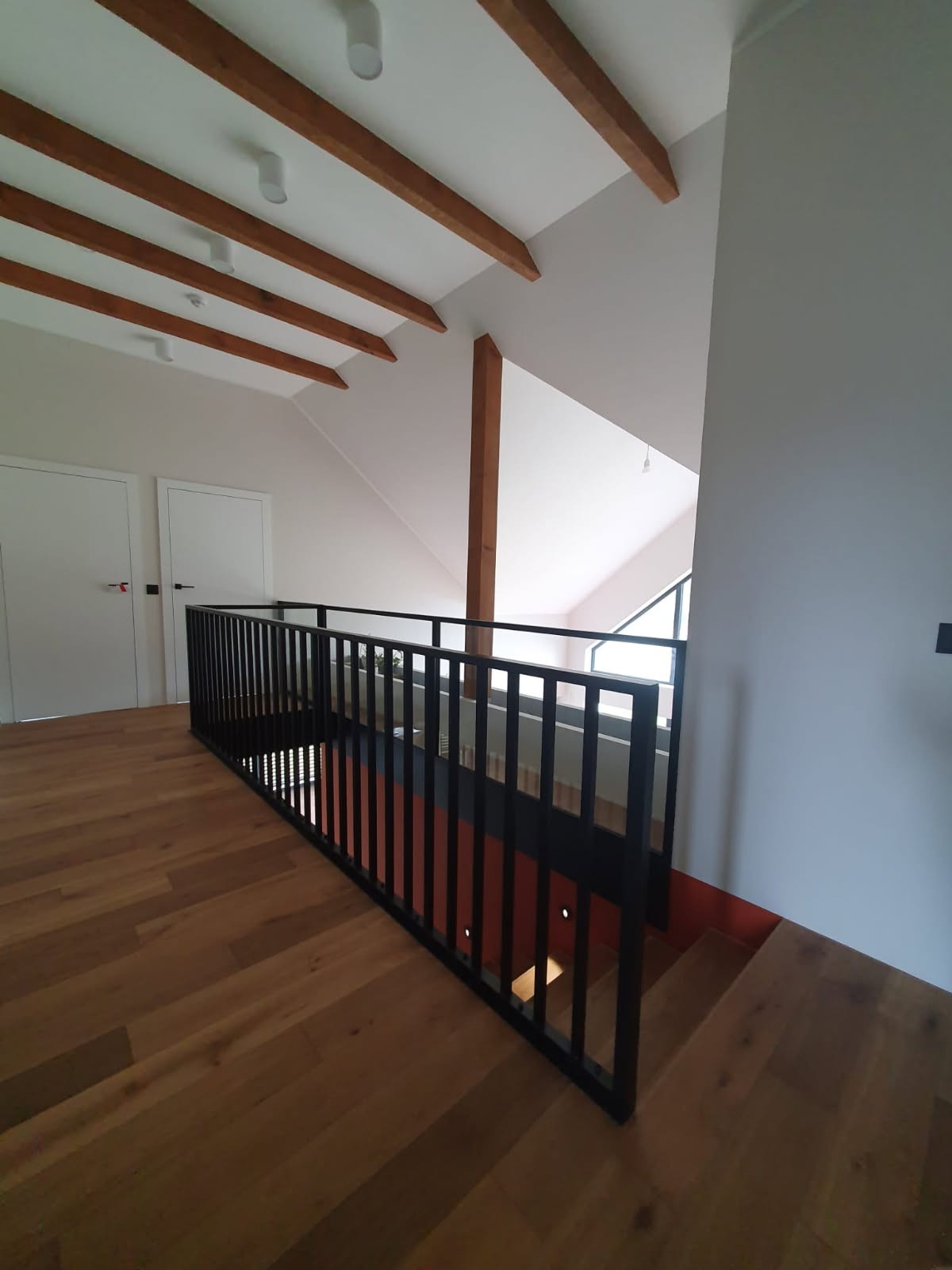 Nowoczesna, czarna metalowa balustrada z pionowymi szczeblami na drewnianym poddaszu z widocznymi belkami stropowymi i białymi drzwiami w tle.