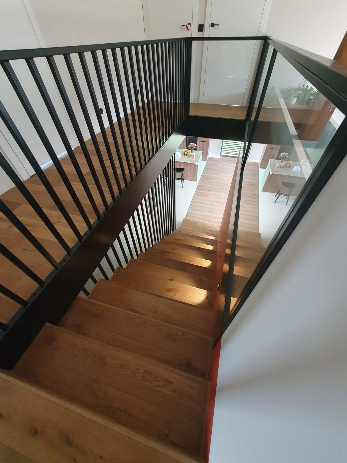 Nowoczesne schody drewniane z czarną metalową balustradą i szklanym panelem bocznym, widok z góry na otwartą przestrzeń kuchni z wyspą.