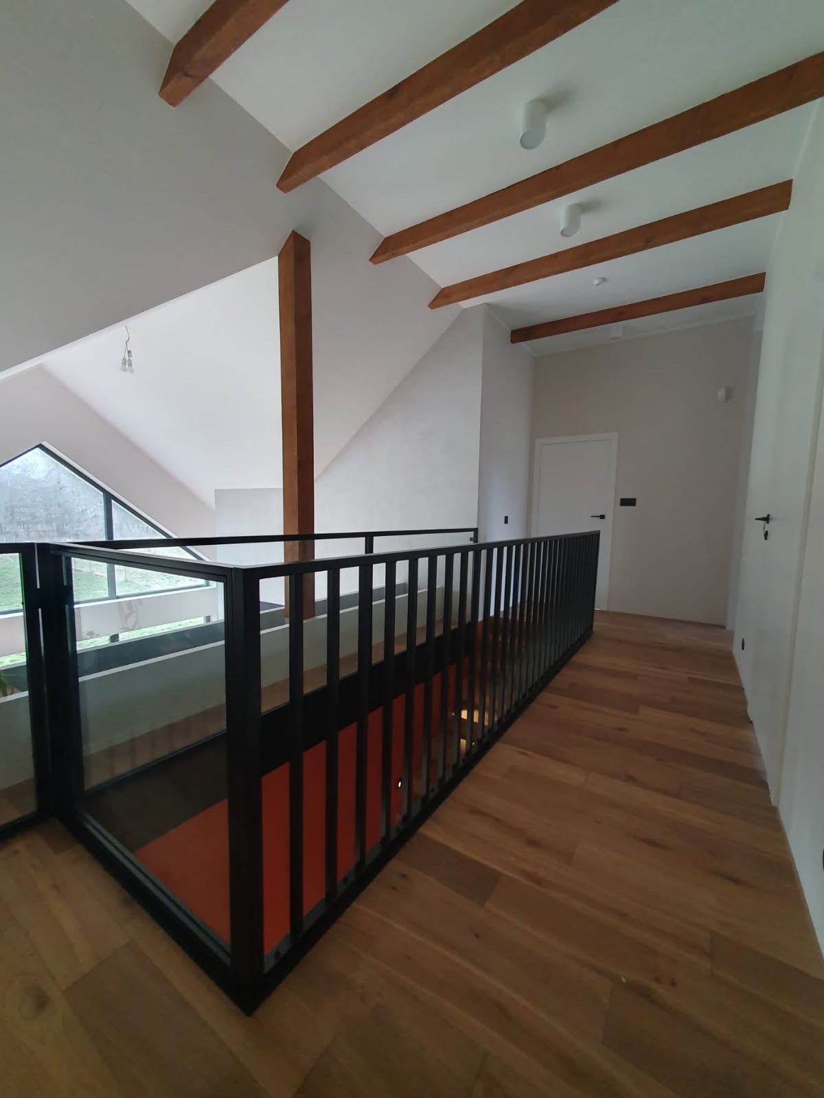 Wewnętrzna balustrada z czarną ramą i szklanym wypełnieniem, drewniana podłoga ułożona w jodełkę, widok na okno i drewniane belki stropowe.