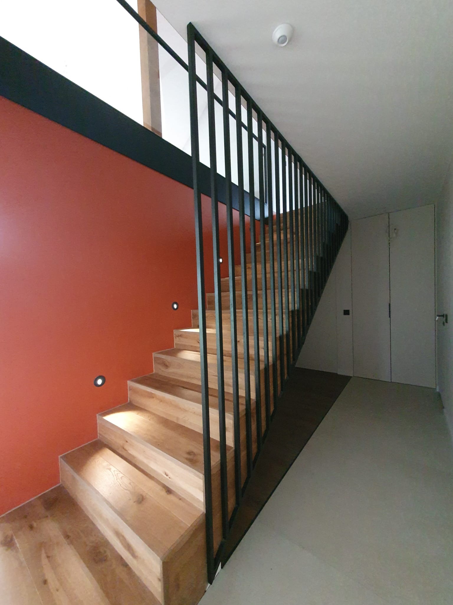 Nowoczesne schody drewniane z czarną, minimalistyczną poręczą z pionowych prętów na tle pomarańczowej ściany z oświetleniem punktowym.