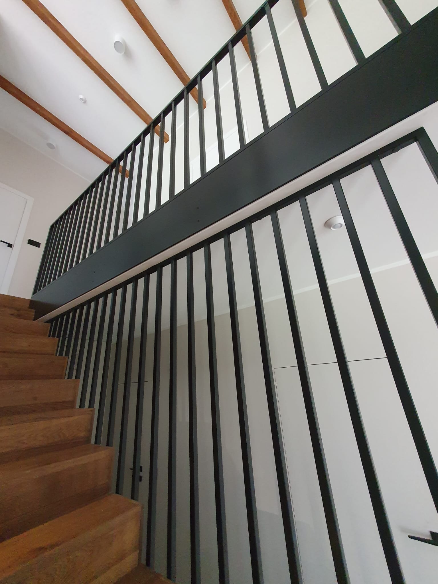 Nowoczesne, czarne poręcze schodowe z pionowymi szczeblami, zamontowane w domu w Śremie, widok z dołu na drewniane schody i białe ściany.