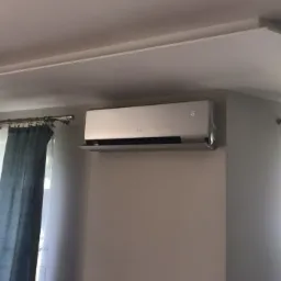 Srebrny klimatyzator ścienny marki Gree, zamontowany w jasnym pokoju z zasłonami w odcieniach szarości i bieli, wyświetlający temperaturę 16 stopni.