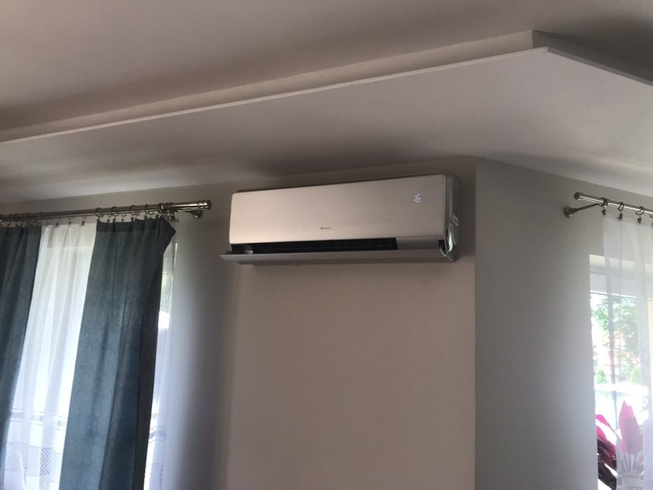 Srebrny klimatyzator ścienny marki Gree, zamontowany w jasnym pokoju z zasłonami w odcieniach szarości i bieli, wyświetlający temperaturę 16 stopni.